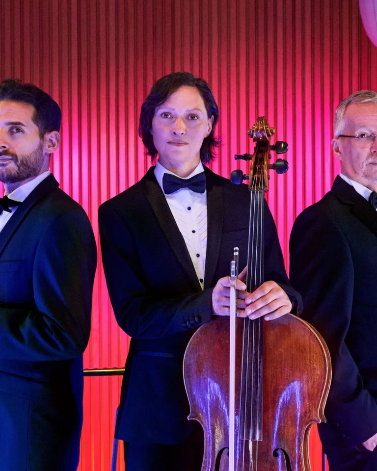Drei Musiker in Anzügen posieren mit ihren Instrumenten: Geige, Cello und Posaune, vor einem rot beleuchteten Hintergrund.