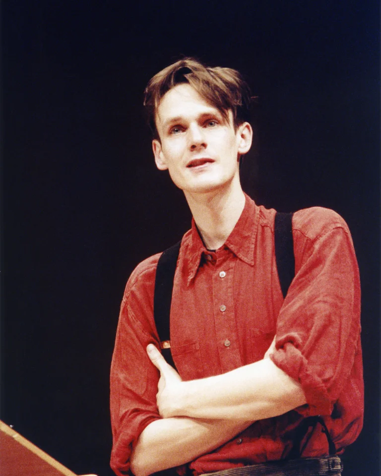 Ein Mann in einem roten Hemd und Hosenträgern steht auf einer Bühne, die Arme verschränkt, vor einem Pult.