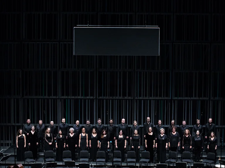 Ein Chor in schwarzer Kleidung (Collegium Vocale Gent) steht in einer Reihe auf einer Bühne mit dunklem Hintergrund für eine Aufführung.