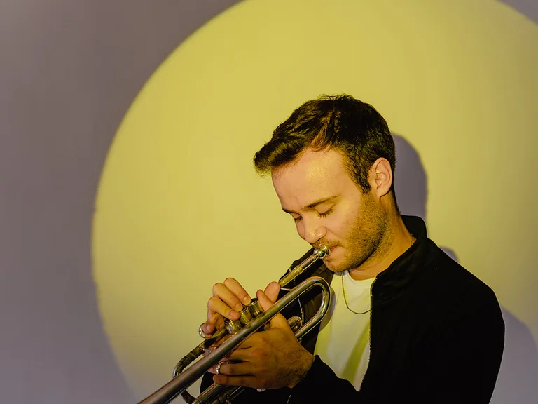 Ein Mann (Simon Höfele) spielt Trompete vor einem gelben Kreis im Hintergrund. Er trägt eine schwarze Jacke und ein weißes T-Shirt.