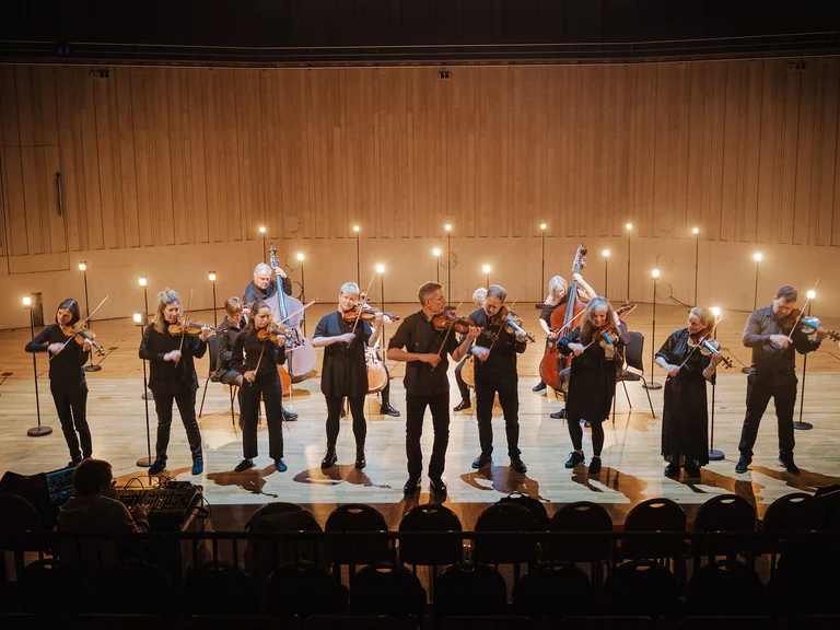 Ein Streichorchester (Scottish Ensemble) auf einer Bühne, das mit Scheinwerfern im Hintergrund spielt. Die Musiker tragen schwarz und halten Geigen.
