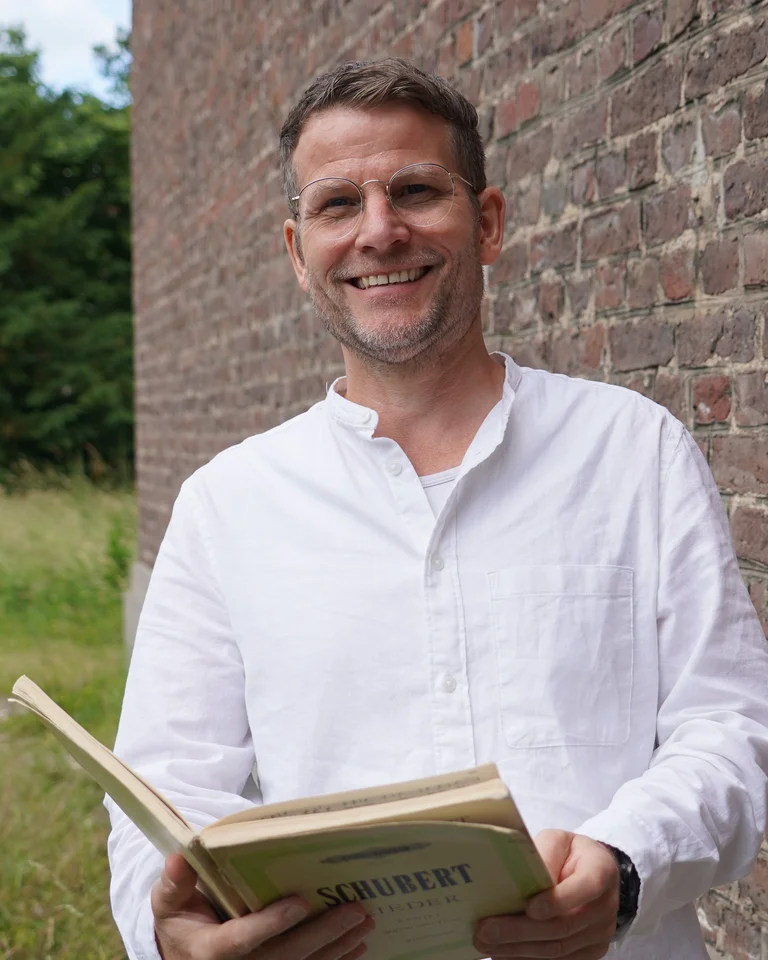 Ein Mann (Jens Lauterbach) in einem weißen Hemd lächelt und hält ein aufgeschlagenes Notenbuch mit der Aufschrift "Schubert" vor einer Ziegelmauer.
