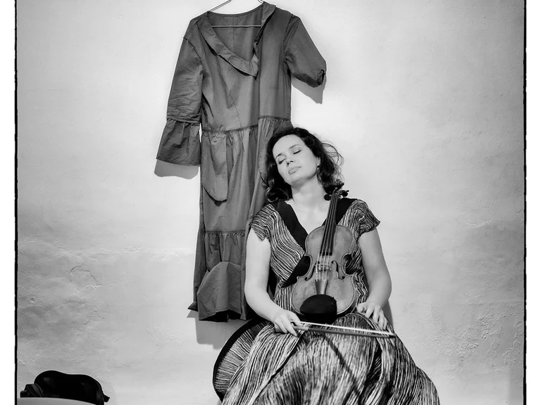 Eine Frau (Patricia Kopatchinskaja) in einem langen Kleid sitzt mit geschlossenen Augen auf einem Stuhl. Über ihr hängt ein zweites Kleid.