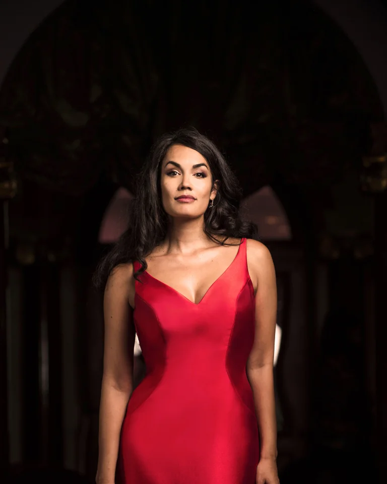 Eine Frau steht in einem eleganten roten Kleid vor dunklem Hintergrund. Sie wirkt selbstbewusst und anmutig.