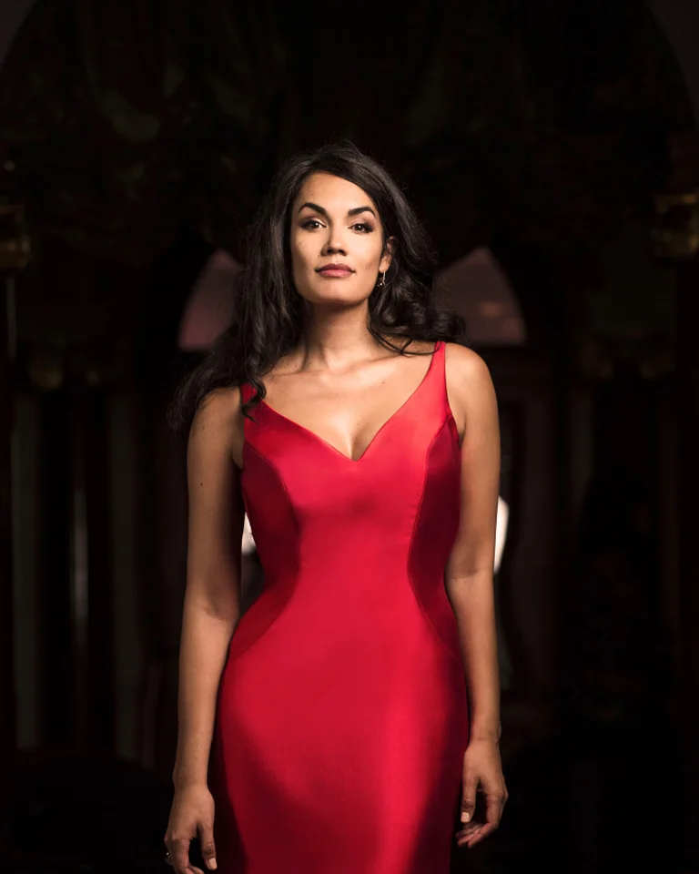 Eine Frau steht in einem eleganten roten Kleid vor dunklem Hintergrund. Sie wirkt selbstbewusst und anmutig.