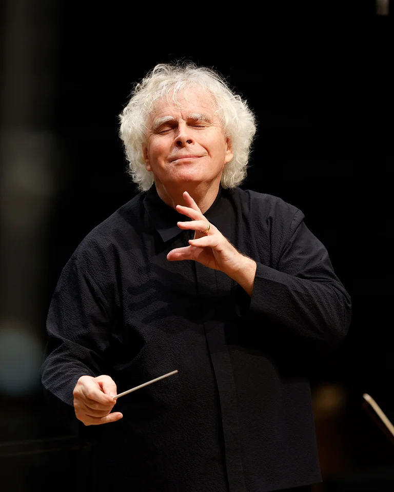 Ein Dirigent (Sir Simon Rattle) führt ein Orchester mit lebhaften Gesten. Er trägt ein schwarzes Hemd und blickt konzentriert.