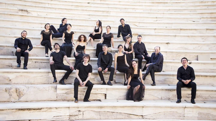 Eine Gruppe von Personen (Il Pomo d'Oro) in schwarzer Kleidung sitzt auf den Stufen eines antiken Theaters im Freien.