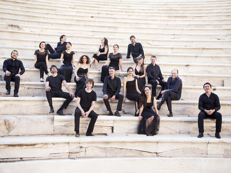 Eine Gruppe von Personen (Il Pomo d'Oro) in schwarzer Kleidung sitzt auf den Stufen eines antiken Theaters im Freien.