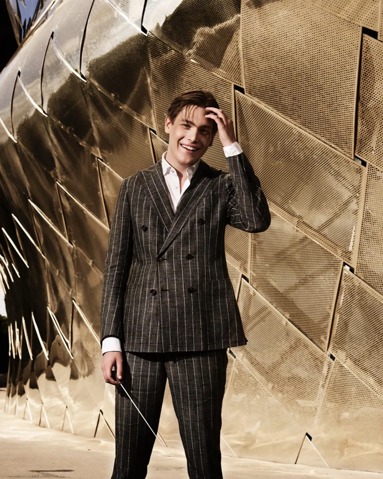 Ein Mann (Klaus Mäkelä) in einem gestreiften Anzug steht vor einer strukturierten, goldenen Wand und lächelt in die Kamera.