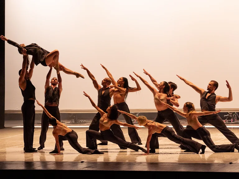 Eine Gruppe von Tänzern auf einer Bühne, die in synchronen Posen eine dynamische Choreografie aufführen. Eine Tänzerin wird hochgehoben.