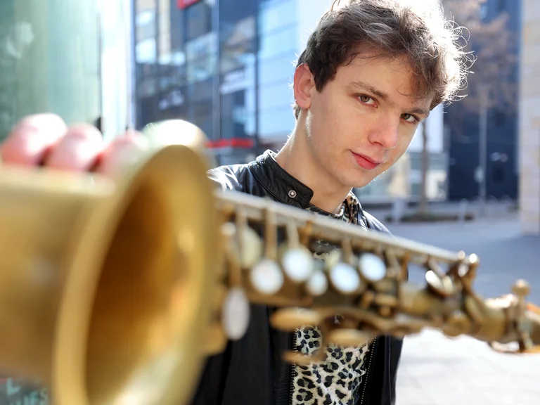 Ein junger Mann hält ein Saxophon vor sich. Er trägt eine schwarze Jacke und schaut ernst in die Kamera.