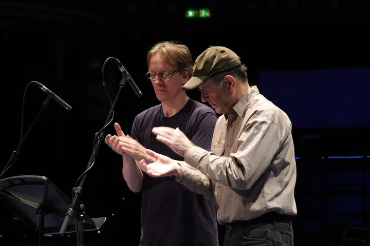 Steve Reich and Rainer Römer (Ensemble Modern) stand in front of microphones and clap their hands.