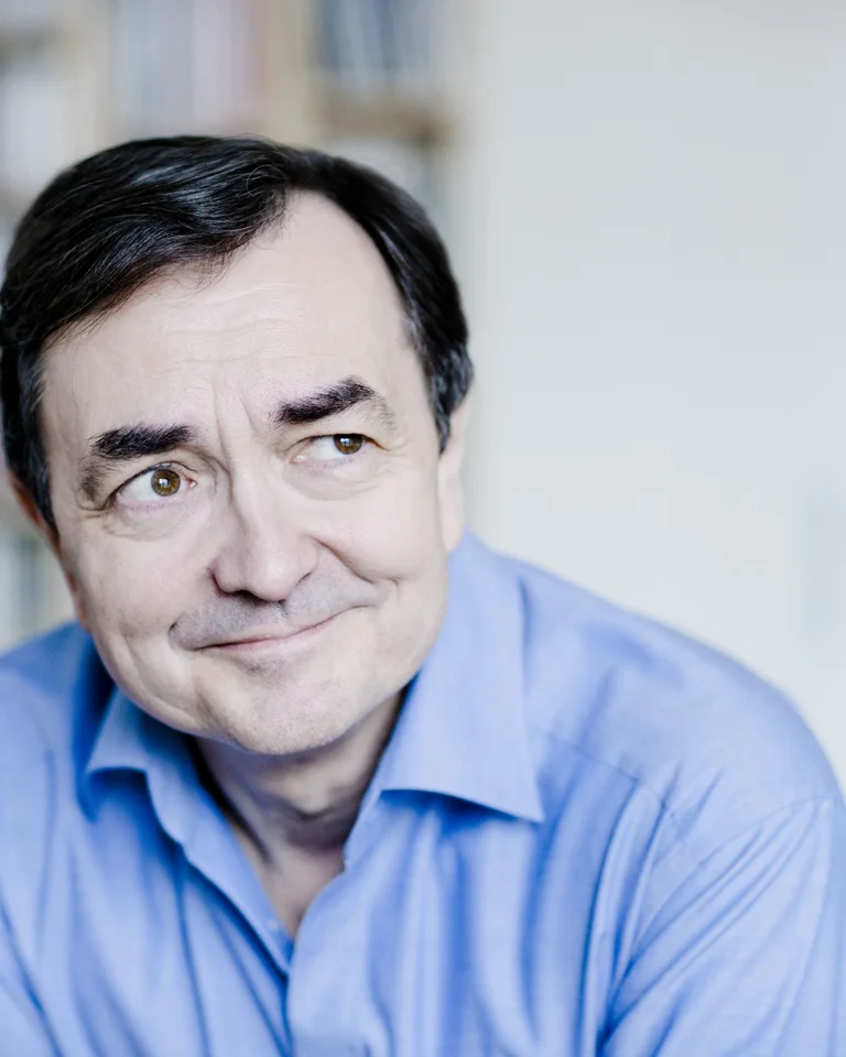 Ein Mann in einem blauen Hemd lächelt sanft. Im Hintergrund sind verschwommene Bücherregale zu sehen.