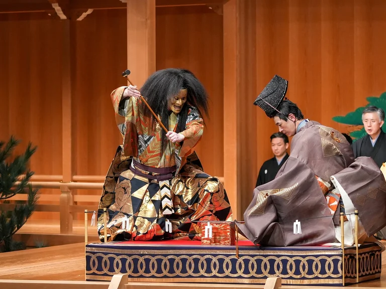 Zwei Künstler führen auf einer traditionellen Noh-Bühne auf. Einer trägt eine dekorative Maske, beide tragen prächtige Kimonos (Kokaji).