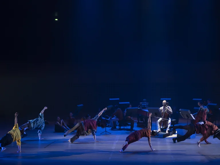 Eine Gruppe von Tänzern in verschiedenen Posen unter blauem Licht auf einer Bühne mit Live-Musikern im Hintergrund.