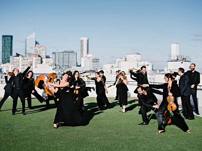 A musical ensemble in formal attire performs on a green rooftop with a city skyline in the background.