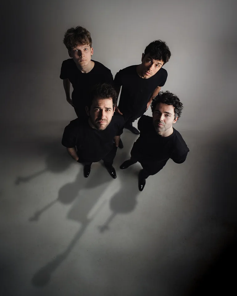 Vier Männer in schwarzer Kleidung (vision string quartet) stehen dicht beieinander, von oben fotografiert. Lange Schatten erstrecken sich über den Boden.