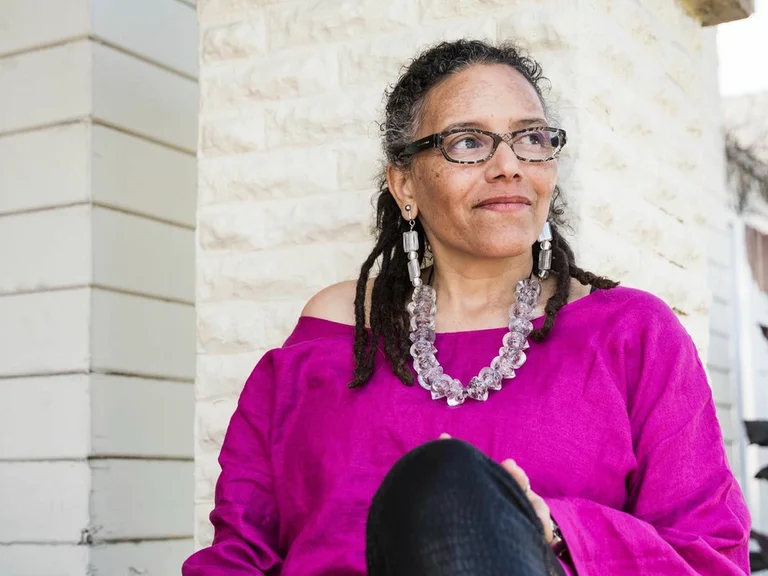 Frau (Nicole Mitchell) sitzt draußen, trägt ein leuchtend pinkes Oberteil und schwarze Hose. Sie hat Brille und Schmuck, schaut nachdenklich.