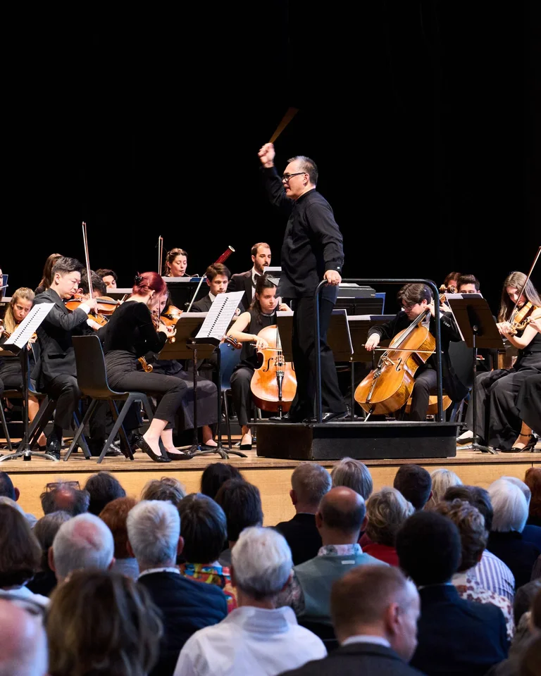 Ein Orchester spielt auf einer Bühne vor einem Publikum, dirigiert von einem Mann mit erhobenem Dirigentenstab.
