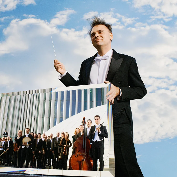 Ein Dirigent im Frack hält einen Taktstock, eine Spiegelung eines Orchesters ist zu sehen, mit Himmel im Hintergrund.