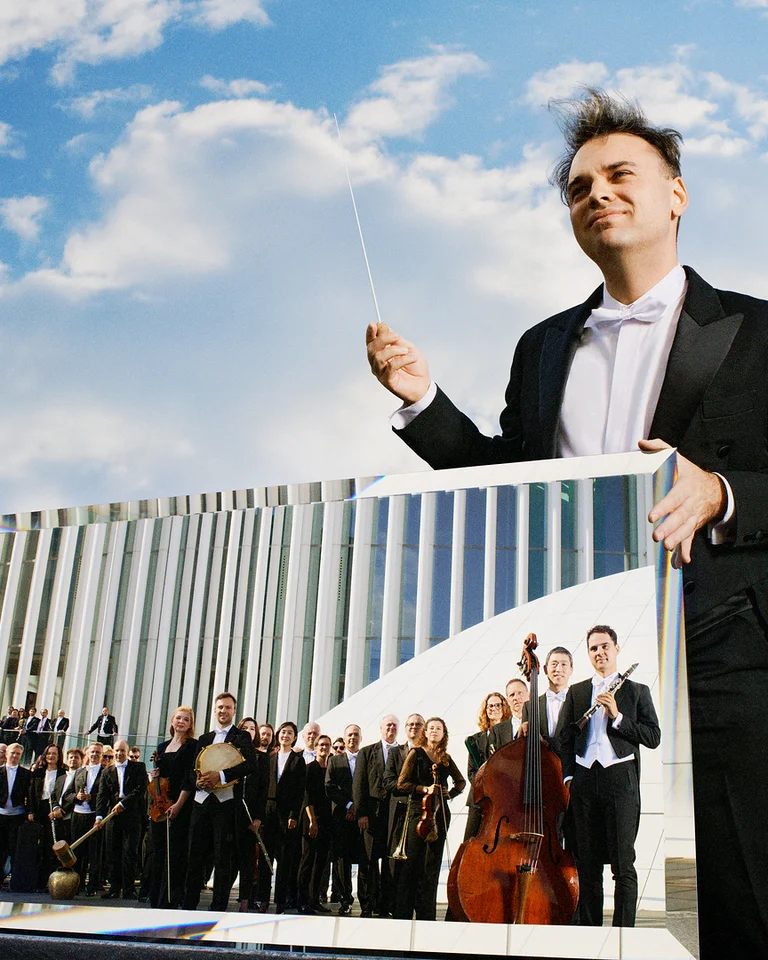 Ein Dirigent im Frack hält einen Taktstock, eine Spiegelung eines Orchesters ist zu sehen, mit Himmel im Hintergrund.