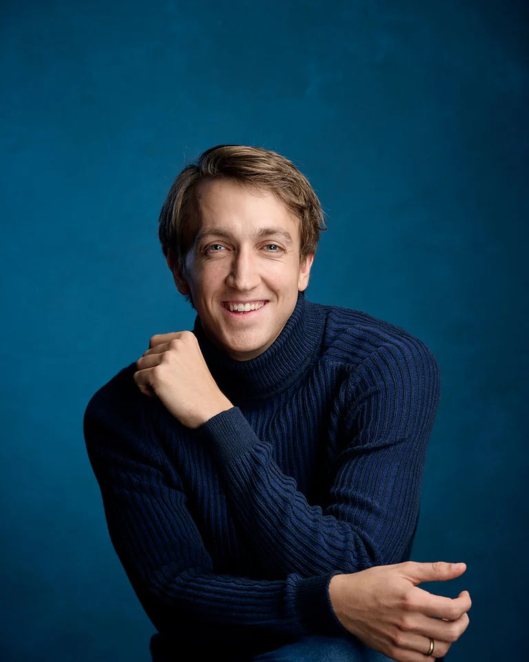 Ein Mann (Fabian Müller) in einem blauen Rollkragenpullover sitzt vor einem blauen Hintergrund. Er lächelt entspannt in die Kamera.