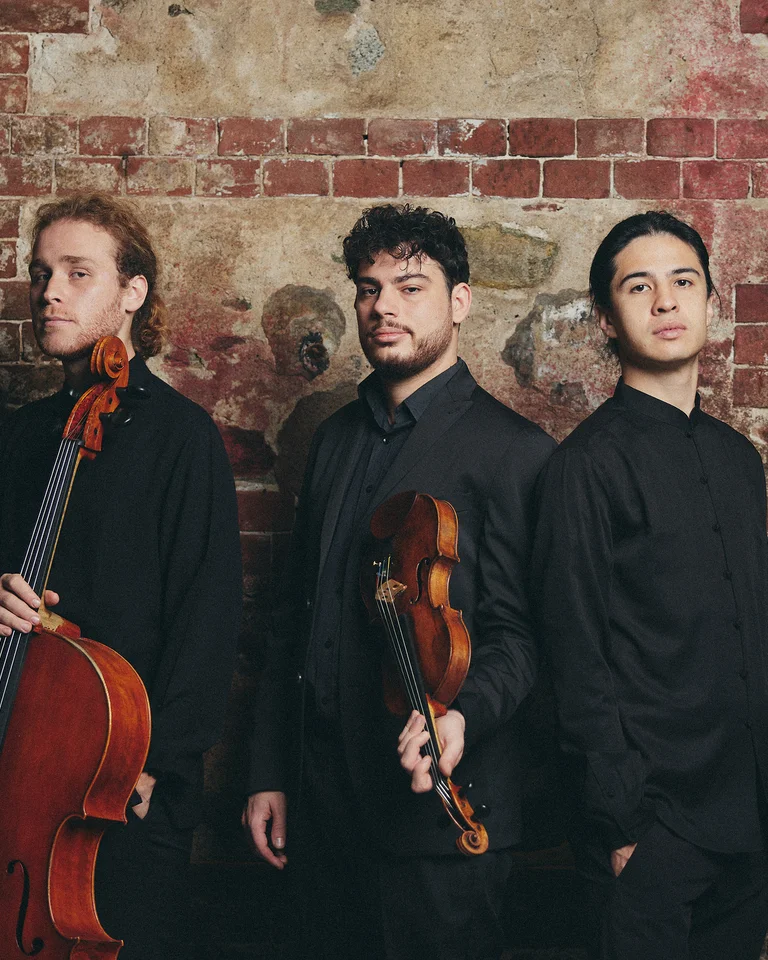 The three members of Trio Concept pose in black attire in front of a brick wall, holding a cello and a violin.