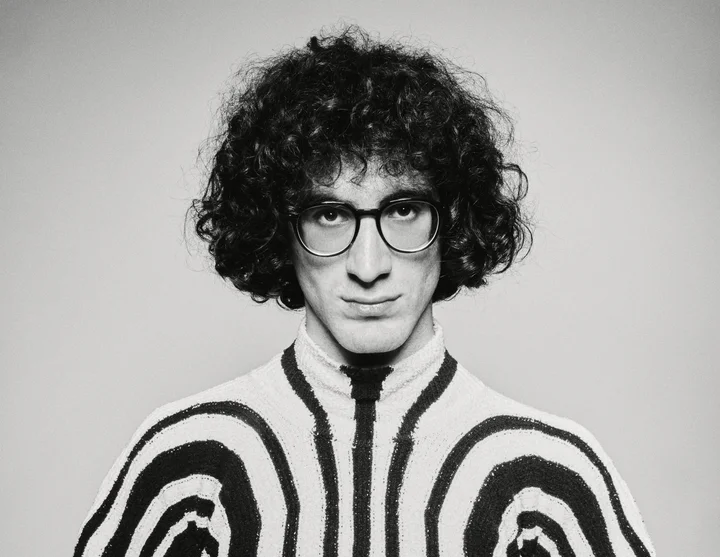 Giorgi Gigashvili schaut mit Brille und lockigem Haar in die Kamera, Er trägt einen schwarz-weißen Pullover mit geschwungenen Streifen.