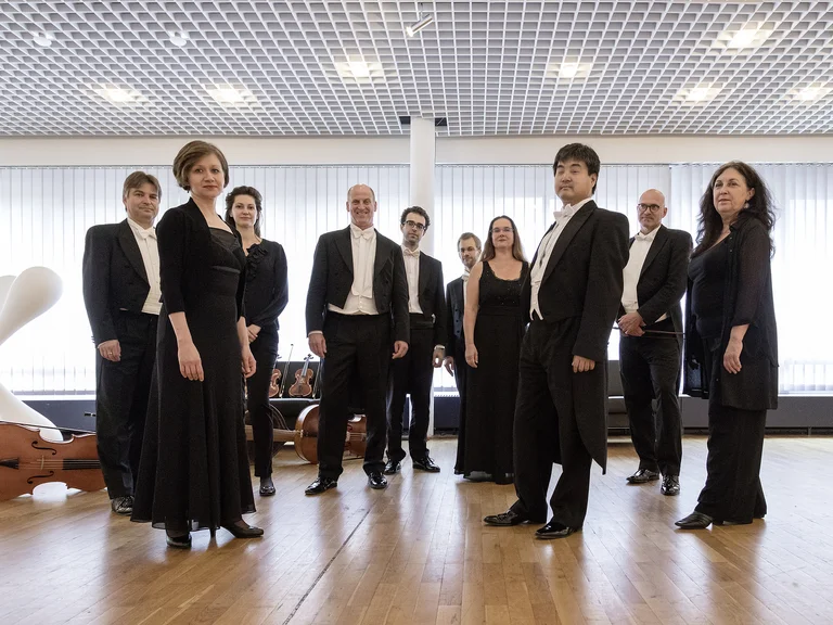 l'arte del mondo: Eine elegante Gruppe von Musikern in formeller Abendkleidung steht in einem hellen Raum mit einem großen Cello im Hintergrund.