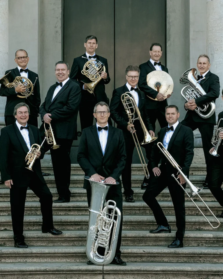 German Brass: A group of musicians in formal suits stands on steps, holding brass instruments like horns and trumpets. They appear poised.
