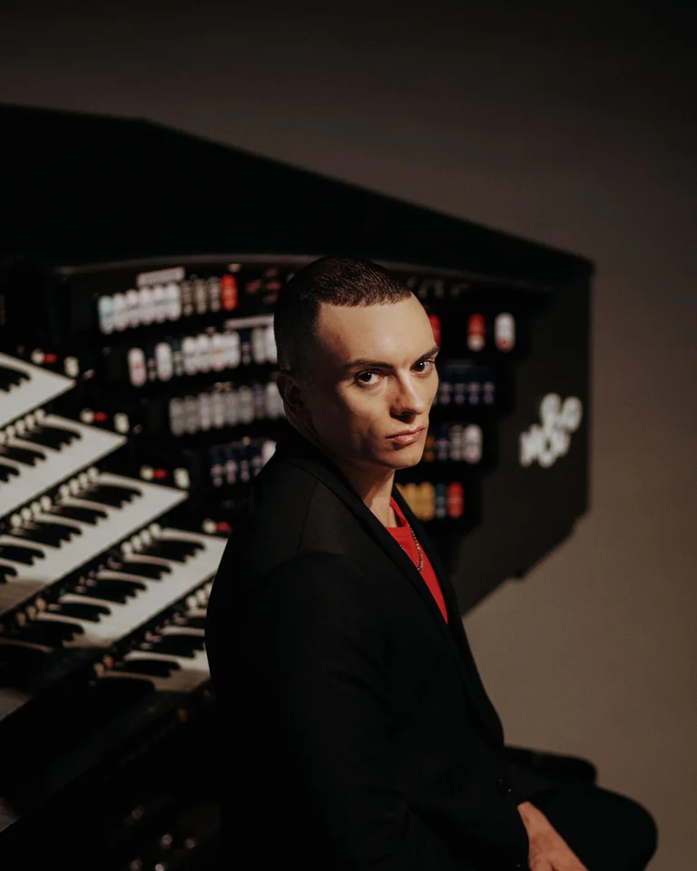 Eine Person sitzt neben einer massiven Orgel mit vielen Tasten und Knöpfen. Die Atmosphäre wirkt kreativ und musikalisch.