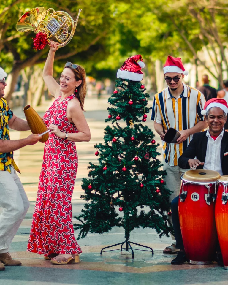 Eine fröhliche Gruppe von Menschen tanzt und musiziert in festlichen Weihnachtsmützen. Sie stehen um einen geschmückten Weihnachtsbaum herum.