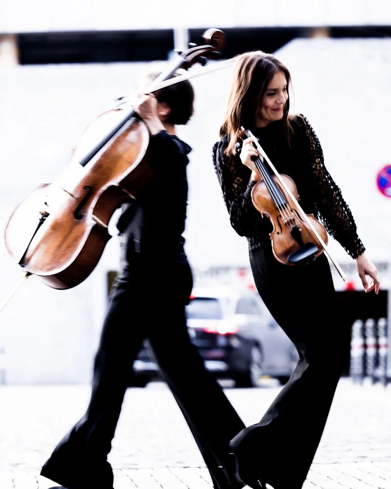 Zwei Musikerinnen gehen lachend eine Straße entlang, eine trägt ein Cello, die andere eine Geige.