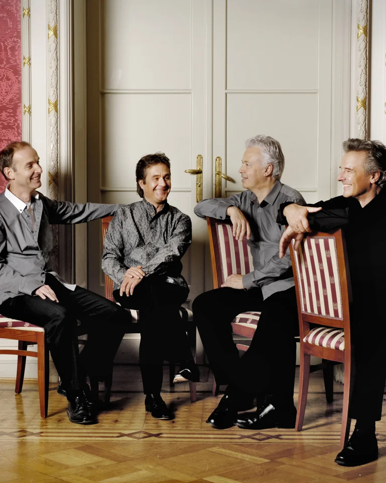 Vier Personen sitzen lächelnd auf Stühlen in einem eleganten Raum mit roten Wänden und Holzparkett.