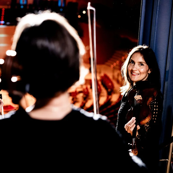 Drei Musiker bereiten sich auf einen Auftritt vor, mit Geigen in der Hand. Eine Frau lächelt in Richtung Kamera.