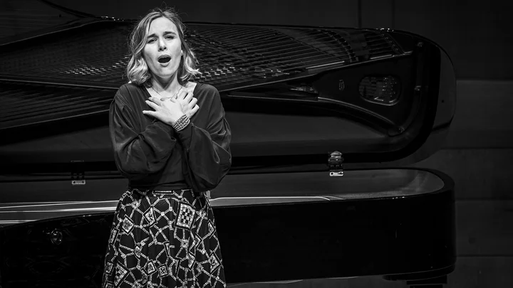 Schwarz-weiß Foto einer Frau, die mit ergreifender Gestik singt. Im Hintergrund ist ein Flügel zu sehen.