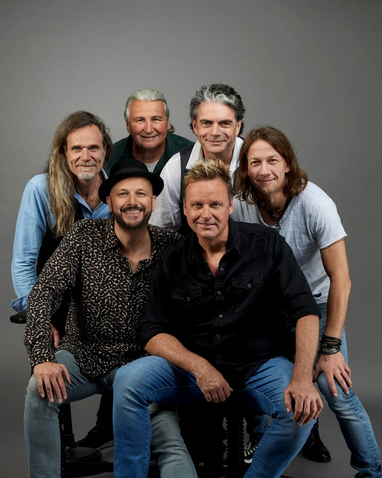 Sechs Männer posieren fröhlich vor einem neutralen Hintergrund, lässig gekleidet. Sie wirken kameradschaftlich und gut gelaunt.