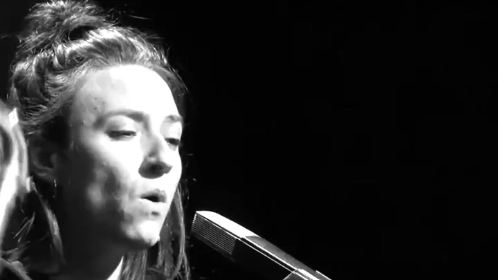 A black-and-white video still of Lucia Cadotsch singing into a microphone with her eyes half-closed.