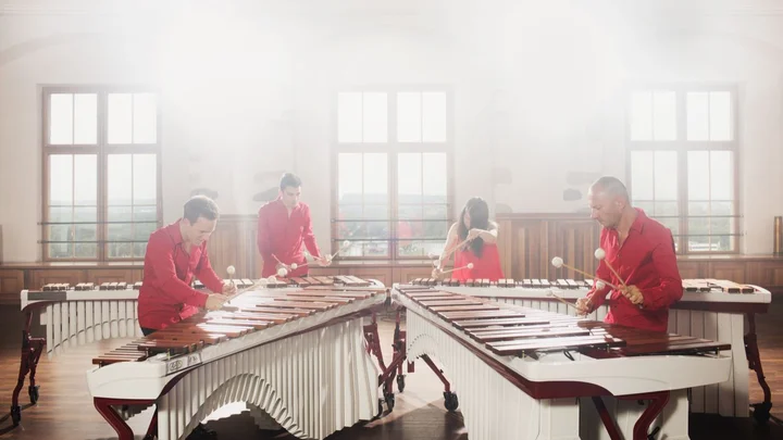 Vier Musiker spielen an zwei großen Marimbaphonen in einem sonnendurchfluteten Raum. Alle sind in rote Anzüge gekleidet.