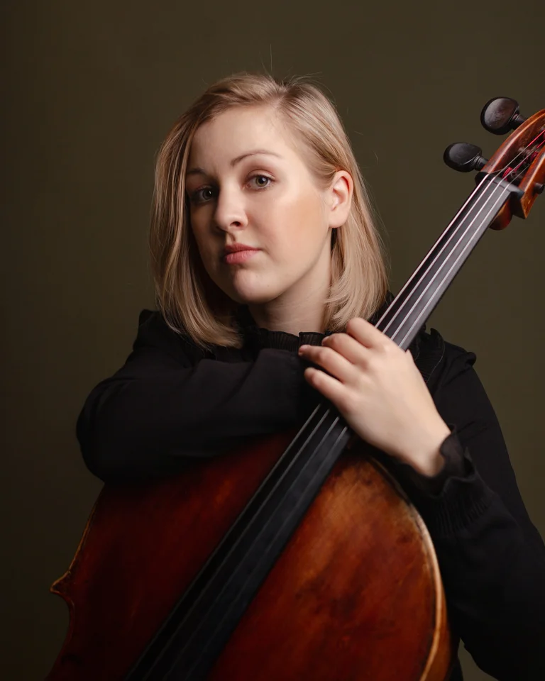 Eine junge Frau mit blonden Haaren hält ein Cello. Sie trägt schwarze Kleidung und schaut direkt in die Kamera.