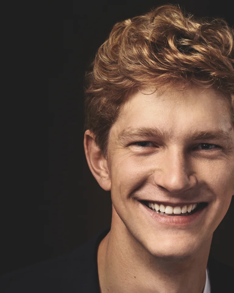 A fotp of Jan Lisiecki: A young man with curly hair smiles warmly against a dark background. He is wearing a black garment.