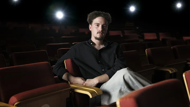 A man sits in an empty theater, illuminated by stage lights, with a serious expression on a red seat.