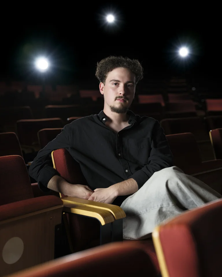 A man sits in an empty theater, illuminated by stage lights, with a serious expression on a red seat.