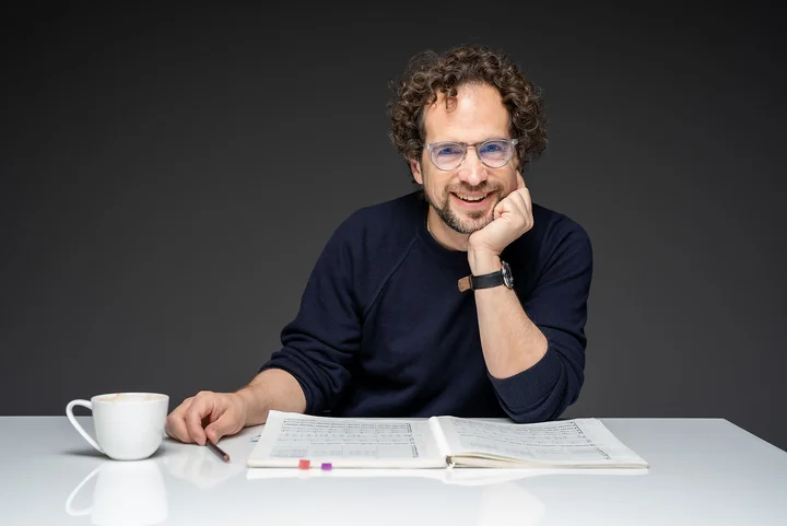 Jonathan Stockhammer sits smiling, his head resting on his left elbow, at a table with a cup of coffee and an open sheet music.