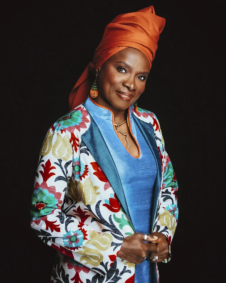 Eine Frau (Angélique Kidjo) trägt ein buntes, florales Jacket über einem blauen Kleid und einen orangefarbenen Kopfschmuck. Sie steht vor einem dunklen Hintergrund.