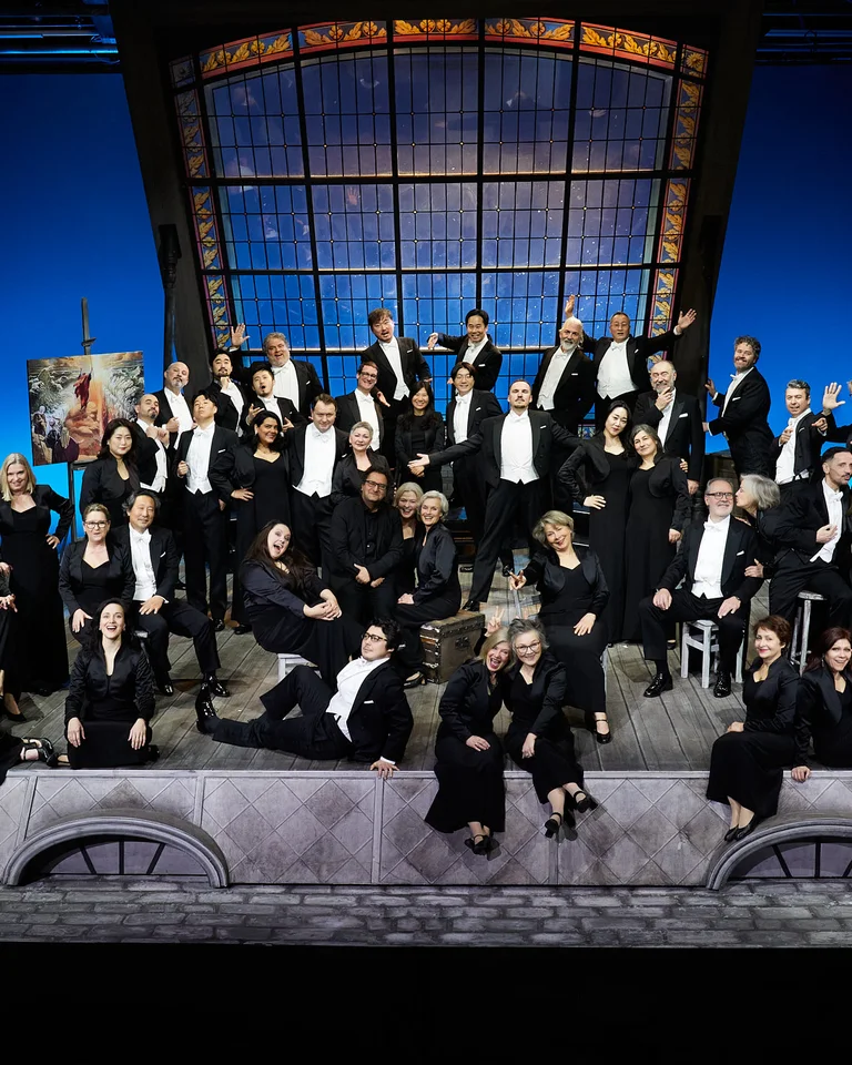 Eine große Gruppe von Menschen in eleganter Kleidung posiert auf einer Bühne mit einem stilvollen, lichtdurchfluteten Hintergrund.