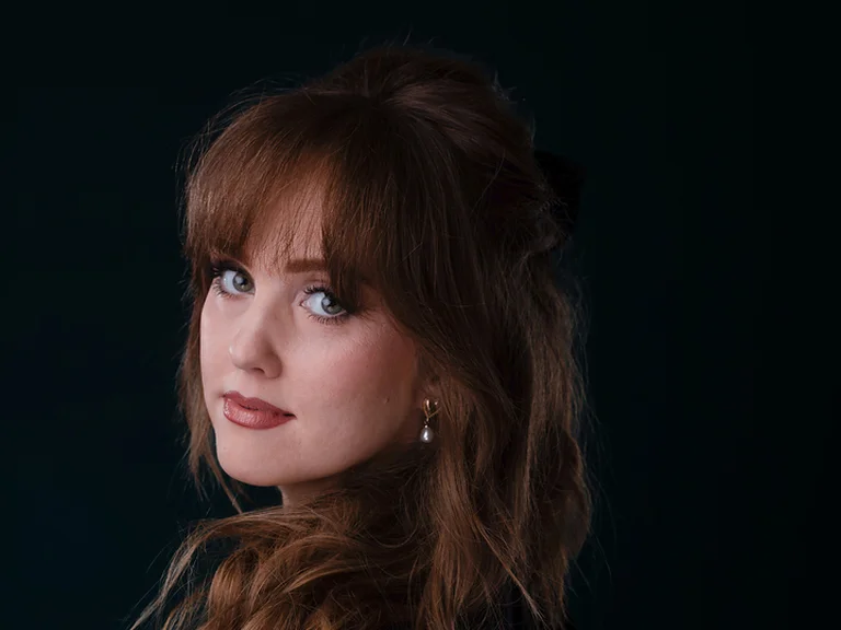 Eine Frau (Mélissa Petit) mit langen, gewellten Haaren und einem sanften Blick, gekleidet in einen schwarzen, halbtransparenten Stoff, vor dunklem Hintergrund.