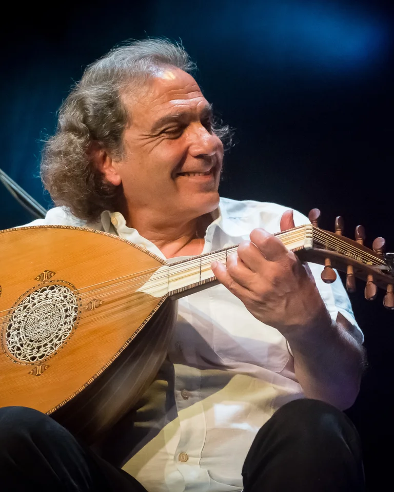 Ein Musiker spielt eine Oud auf der Bühne. Er trägt ein weißes Hemd und blickt lächelnd zur Seite. Ein Mikrofon ist sichtbar.