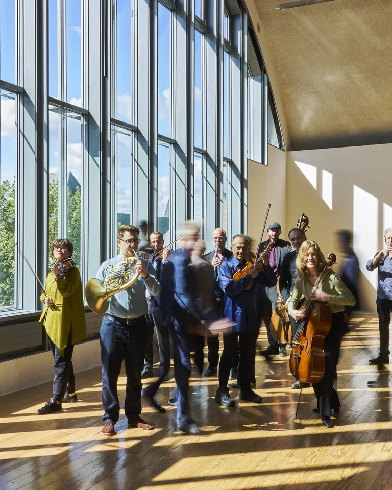 Die Mitglieder des Ensemble Modern, darunter einige Musiker:innen mit Instrumenten, stehen und bewegen sich in einem lichtdurchfluteten Raum mit großen Fenstern und Holzboden.