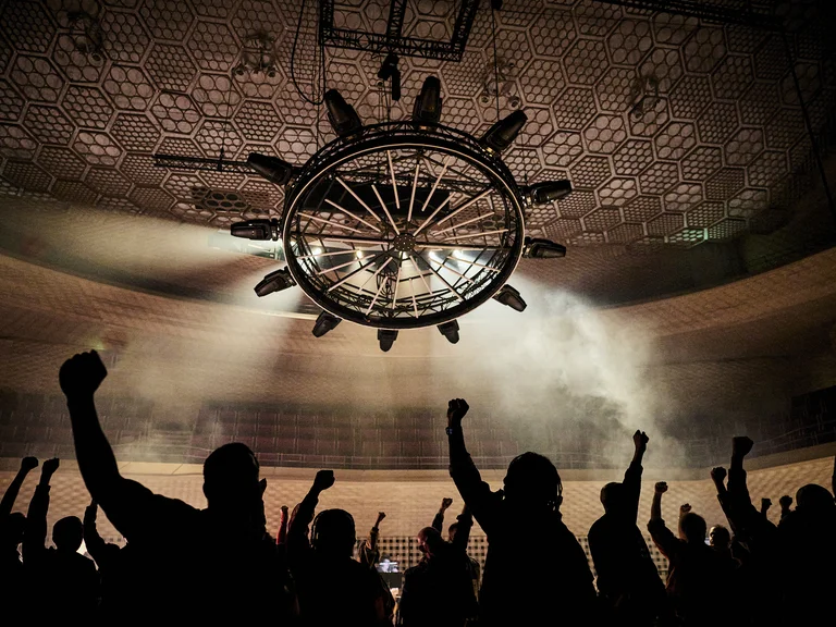 Silhouettes of people (Structure Rualitè mit Capella Mediterranea u.a.) with raised fists in a lit hall. A large, ornate ceiling light hangs above them.